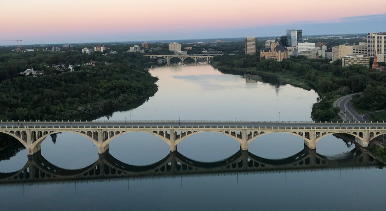 Aerial view of a city bridge reflecting over a calm river at sunset, representing our community in Saskatoon
